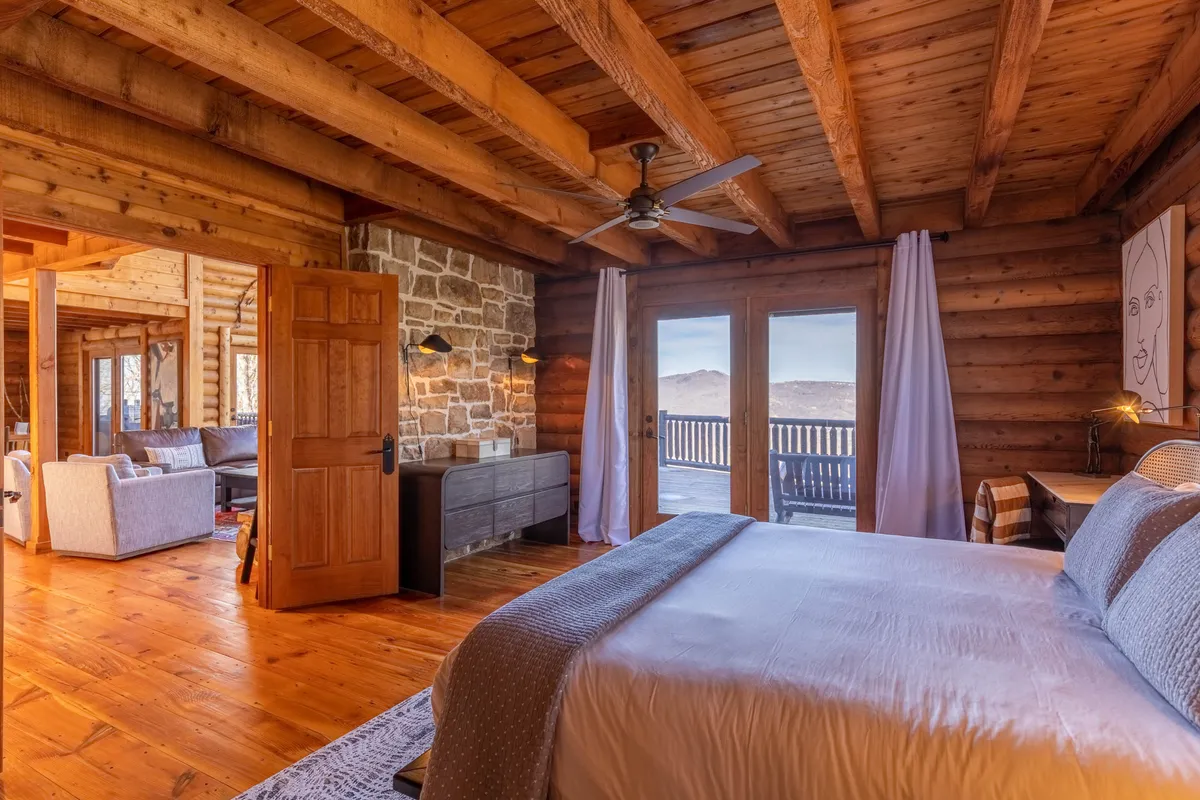 A spacious bedroom with large windows offering mountain views, wooden beams, a stone wall, and a bed adorned with decorative pillows. The setting is warm and sophisticated, with touches of rustic elegance and an inviting feel.