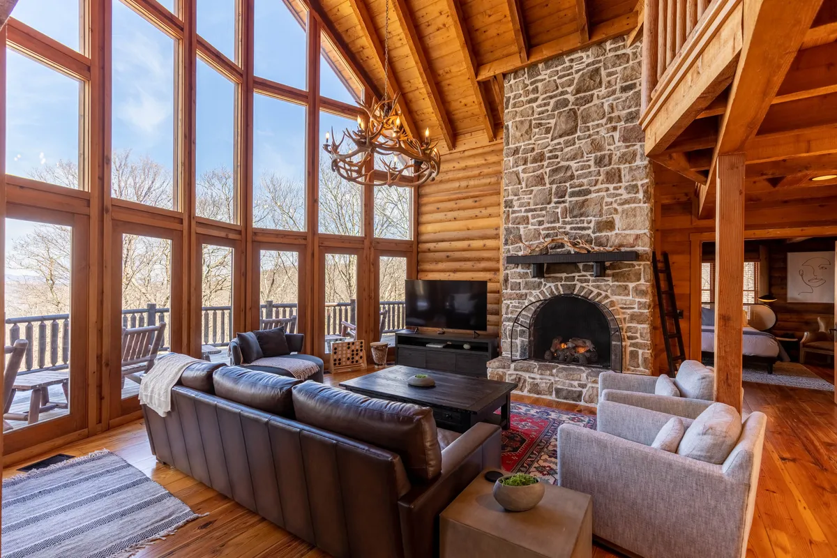 A welcoming living room with large windows allowing natural light to pour in, a striking stone fireplace as the focal point, a chandelier above, and comfortable sofas arranged for conversation and relaxation. The view from the windows hints at the scenic setting, while the space feels ideal for both intimate gatherings and larger family moments.