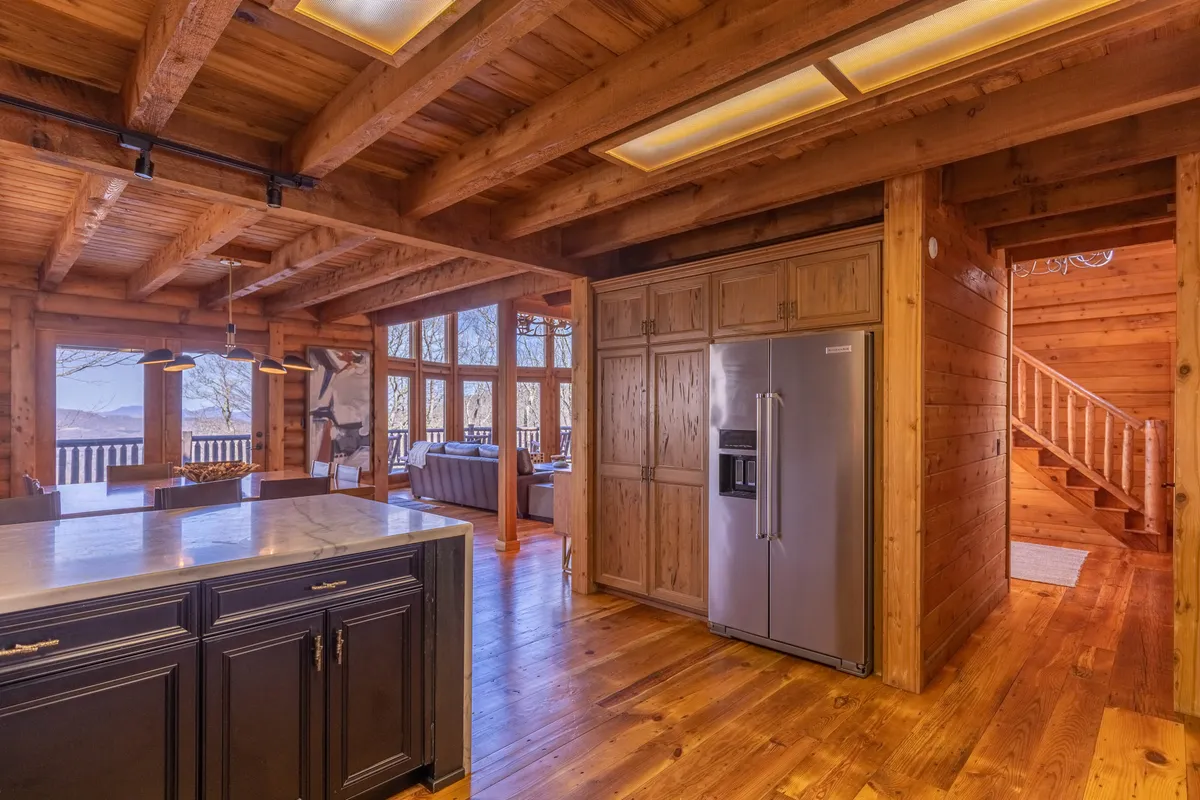 A wide view of the kitchen featuring an expansive marble island with stylish black cabinetry, a stainless steel refrigerator, exposed wooden beams, and large windows framing mountain vistas. The space feels open, bright, and perfect for group meal prep or sharing a morning coffee.