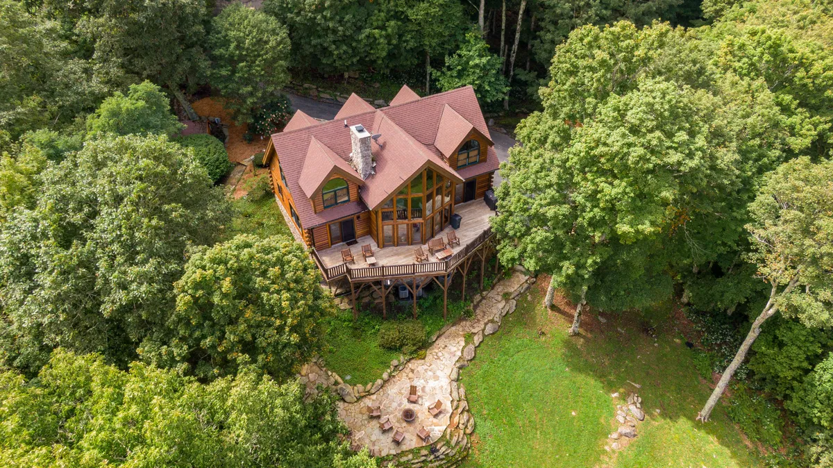 The image showcases a stunning log cabin nestled within a quiet forest, with a spacious deck, inviting fire pit, and panoramic mountain views visible in the background. The setting exudes tranquility and offers ample space for outdoor gatherings, relaxation, and taking in the sunset.