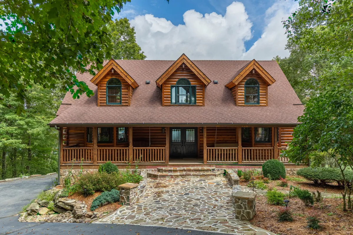 A picturesque exterior shot of the log cabin nestled amongst greenery, featuring a welcoming stone pathway leading up to a front porch surrounded by a landscaped garden. The image evokes a sense of arrival, privacy, and connection to nature, with outdoor space for gatherings and relaxation.