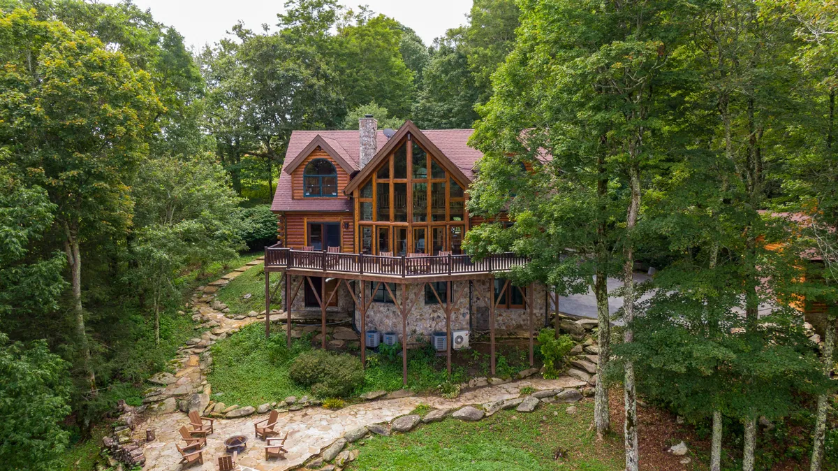 A large wooden deck and stone patio extend from the log cabin exterior, nestled among tall trees. The space is furnished for gatherings, with a fire pit in view, and the serene forest surroundings evoke a tranquil, private atmosphere ideal for team-building or relaxation.