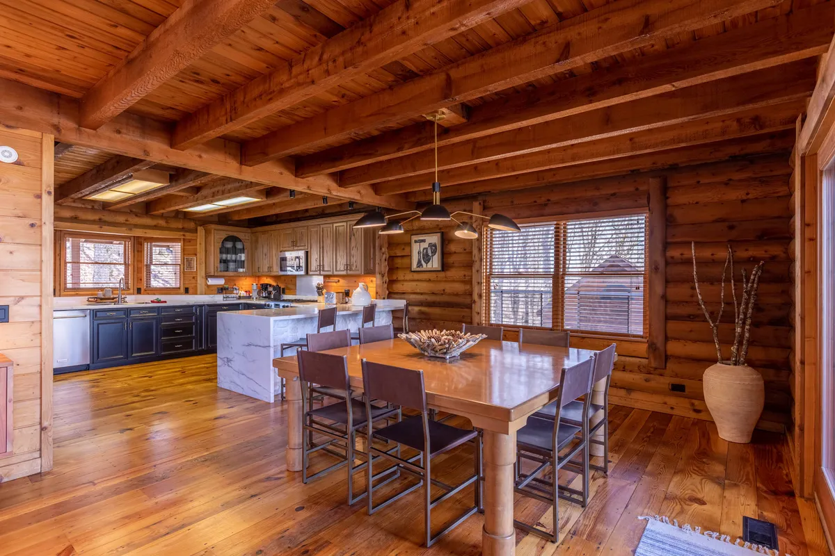 A seamless transition from the kitchen to the dining area is on display here. The image shows a dining table set near a kitchen island, with rustic wooden beams and large windows spilling light onto the communal area—ideal for both cooking and shared dining experiences.