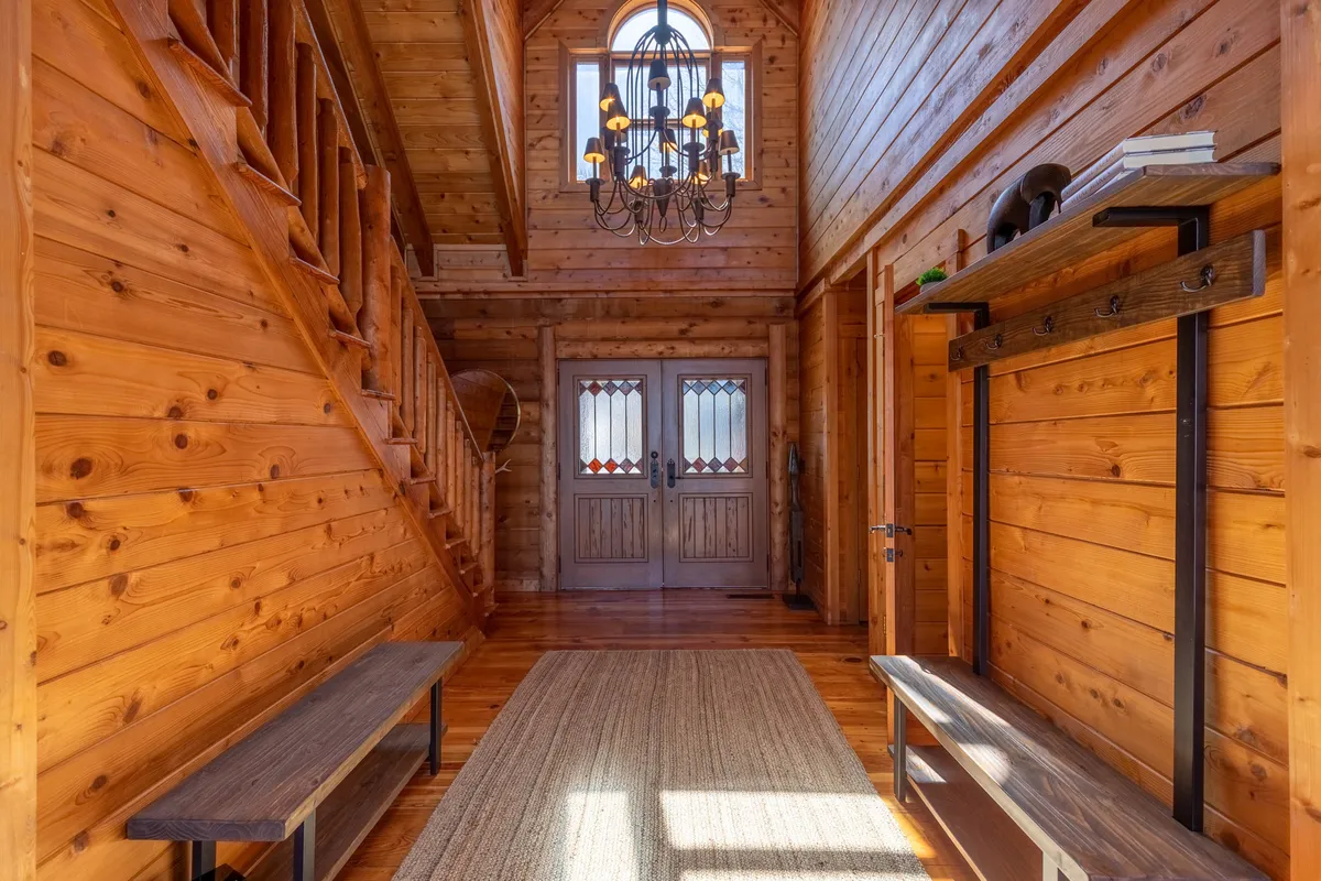 A warmly lit entryway featuring a wooden staircase, double doors, and rustic wooden benches. The space exudes a welcoming first impression with its natural wood tones and inviting decor, setting the stage for the rest of the home.