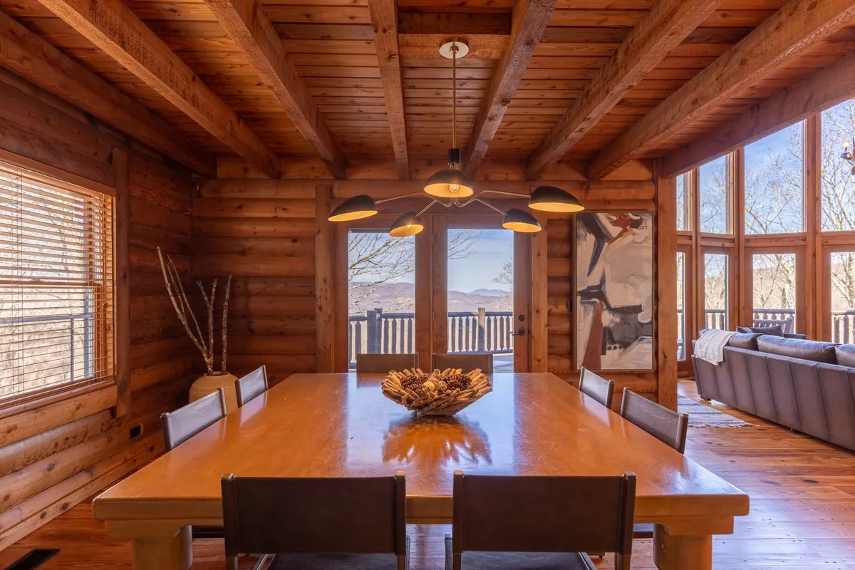 A cozy dining table adorned with a simple centerpiece sits near expansive windows offering scenic views. The modern light fixture and abundant natural light create a warm, inviting spot for meals, conversations, or celebrations.