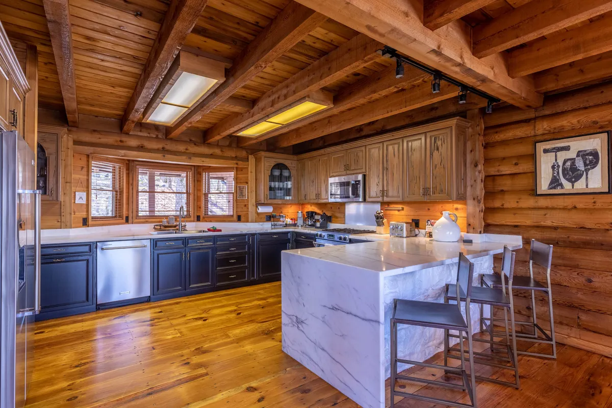 This image spotlights a striking marble kitchen island that echoes the 'marble waterfall peninsula' described in the section. The scene is accented by wooden beams overhead, sleek black cabinetry, expansive windows, and a harmonious blend of contemporary and rustic elements. Appliances are visible, indicating a fully equipped kitchen ready for gourmet cooking or casual gatherings.