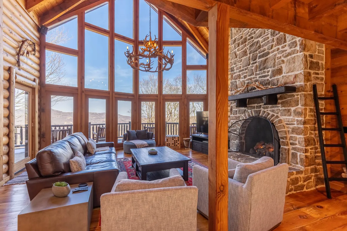 A spacious interior with large panoramic windows framing mountain views, a stone fireplace, exposed wooden beams, and plush seating. The setting is both comfortable and visually connected to the outdoors, blending interior coziness with the natural landscape.