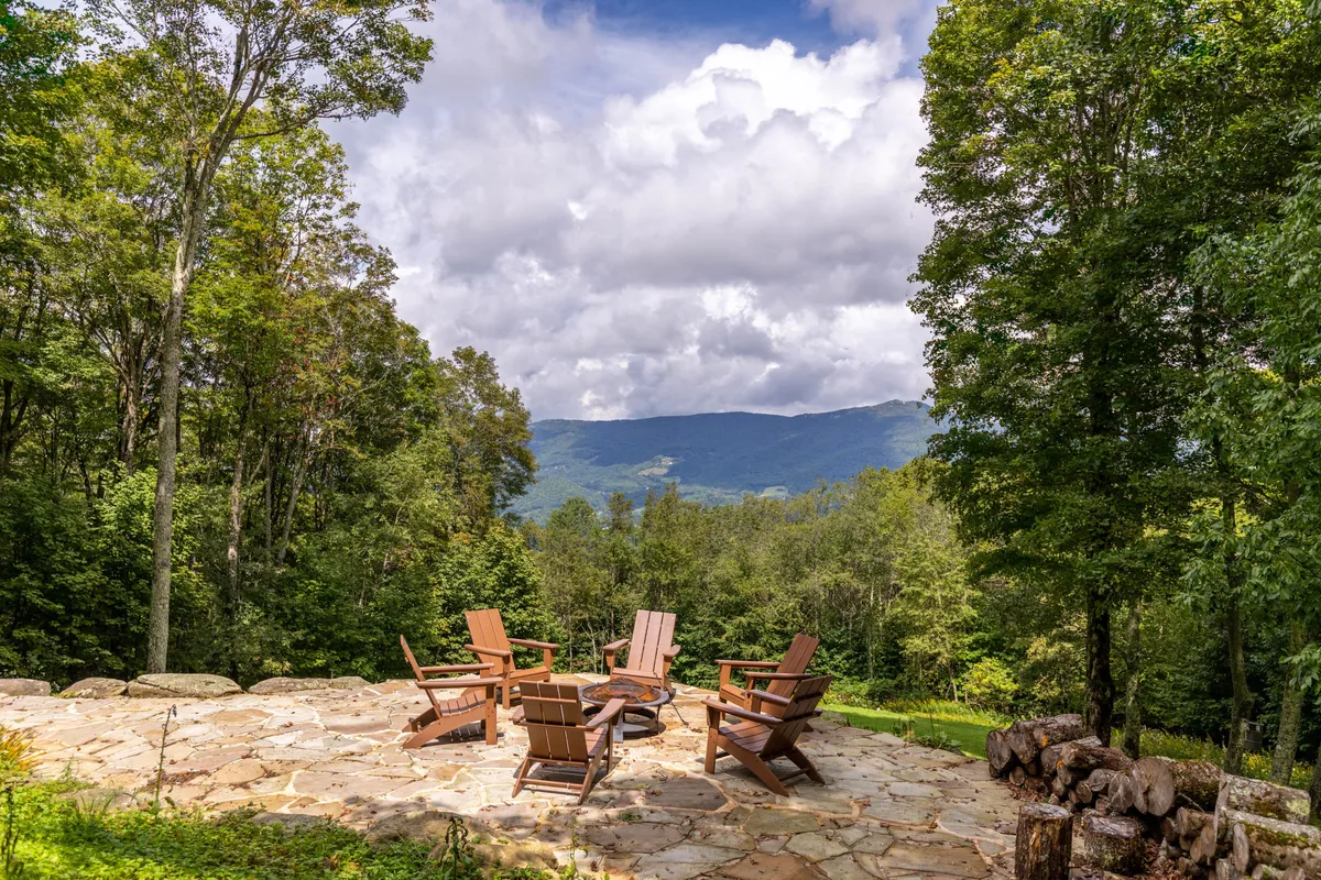 An inviting fire pit area surrounded by wooden chairs and trees, with mountain views peeking through in the background. This serene communal outdoor space is ideal for evening gatherings, storytelling, or enjoying sunset vistas after a day of local exploration.