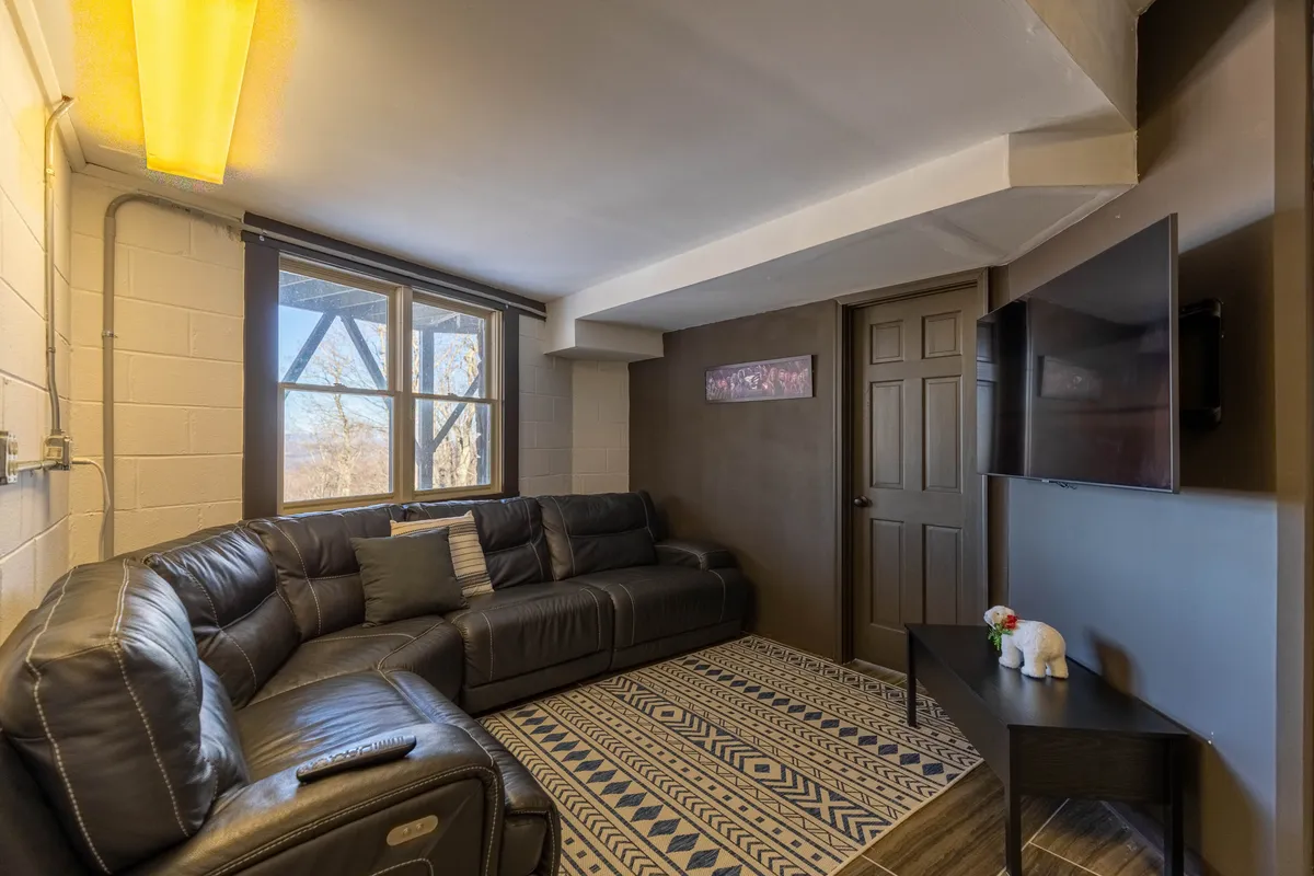 A cozy lounge area with a leather sectional couch facing a large window and a prominent television, set atop a decorative rug. The arrangement is designed for comfort and modern entertainment, ideal for movie marathons or quiet moments with a book.
